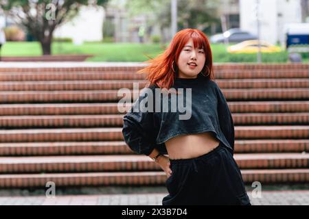 Femme coréenne aux cheveux roux frappant une pose de danse sur les marches urbaines. Banque D'Images