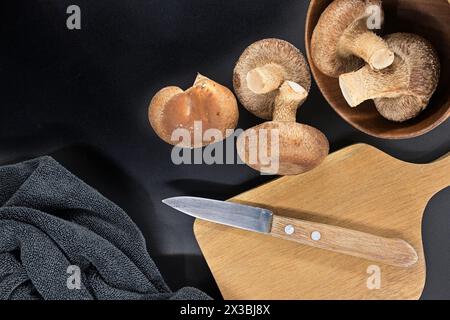 Bol contenant des champignons shiitake frais, planche à découper en bois avec couteau dessus et serviette sombre disposée sur fond noir Banque D'Images