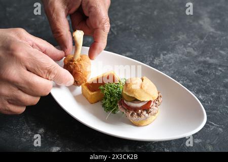 Mains organiser un cours de restauration rapide gastronomique d'un mini hamburger, carré de pommes de terre frites avec ketchup et un pilon de poulet sur une assiette blanche, espace copie, sele Banque D'Images