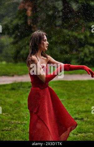 Une jeune femme dans une robe rouge fluide et de longs gants danse gracieusement sous la pluie d'été, incarnant l'équilibre et l'élégance. Banque D'Images