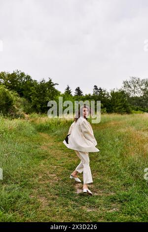 Une belle jeune femme dans une robe blanche marchant gracieusement à travers un champ luxuriant, se prélasser dans la brise d'été. Banque D'Images