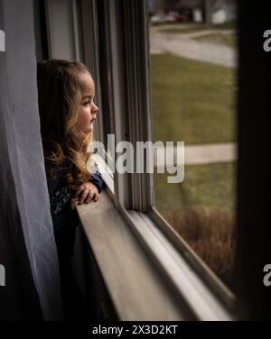 Belle petite fille avec des boucles blondes regardant par la fenêtre en été Banque D'Images