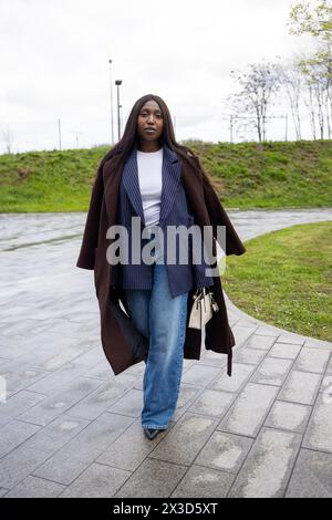 Cette photo captivante représente une jeune femme noire se promenant vigoureusement dans un parc urbain. Elle a un ensemble d'automne à plusieurs couches, avec un superbe blazer à fines rayures sous un manteau brun ouvert et riche. Son haut blanc, son Jean bleu classique et son sac à main élégant complètent le look, incarnant un mélange de praticité et de sensibilité à la mode. Dynamic Urban Walk : femme avant-gardiste en vêtements d'automne superposés. Photo de haute qualité Banque D'Images