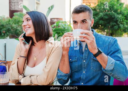 Un bel homme boit et profite de sa tasse de café sur la terrasse d'un restaurant tandis que sa petite amie a une conversation téléphonique. Jeune couple adulte caucasien reposant ensemble sur un coffeeshop. Photo de haute qualité Banque D'Images
