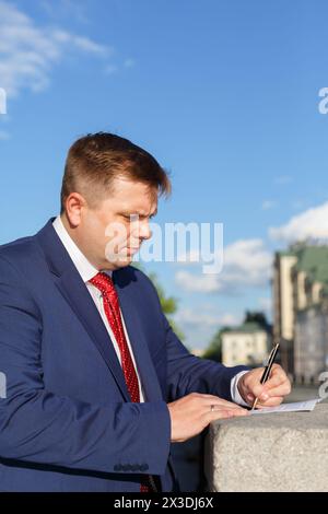 portrait de demi-croissance de beau jeune homme d'affaires en costume à l'extérieur, signe des documents Banque D'Images