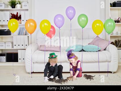 deux enfants jouant avec des chatons sur fond de chambre avec un grand canapé blanc doux, tapis léger sur le sol et des ballons de rangée suspendus dans l'air Banque D'Images