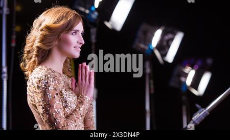Portrait d'une femme heureuse en robe brillante avec reliées les unes aux autres paumes devant des projecteurs dans l'obscurité Banque D'Images