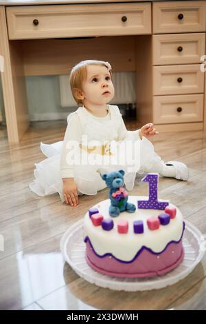 Petite fille en robe blanche se trouve sur le sol près du gâteau d'anniversaire décoré avec ours et numéro un. Banque D'Images