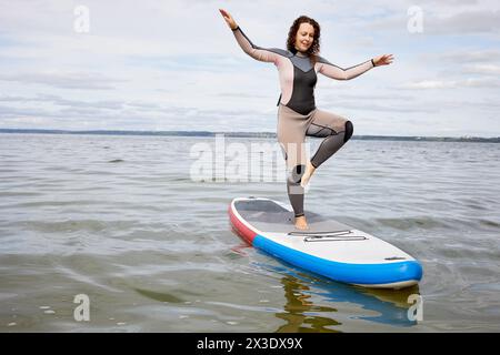 Jeune femme équilibre sur une jambe sur le panneau gonflable SUP sur l'eau. Banque D'Images