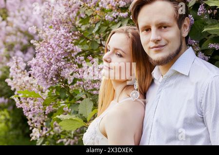 Jeune homme barbu et femme se tiennent ensemble dans le parc près du buisson fleuri de lilas. Banque D'Images
