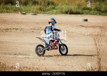 Garçon en costume et casque monte moto sportive sur route de terre poussiéreuse. Banque D'Images