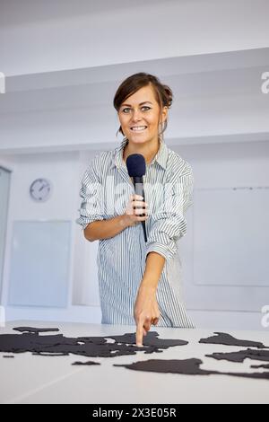 Jeune femme souriante avec microphone pointe doigt sur des fragments de carte du monde sur le bureau. Banque D'Images