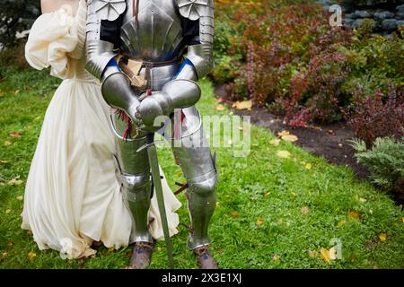 Homme habillé en costume d'armure de chevalier avec épée et fille dans la robe magnifique stand en plein air sur l'herbe. Banque D'Images