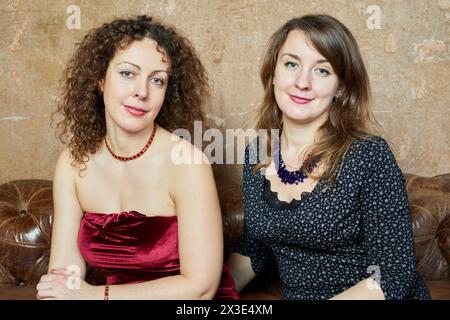 Portrait de deux femmes assises sur un vieux canapé en cuir dans une chambre avec des murs déchiquetés. Banque D'Images