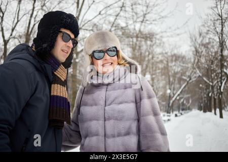 Souriant homme et femme dans des lunettes de soleil sur l'allée dans le parc d'hiver enneigé. Banque D'Images