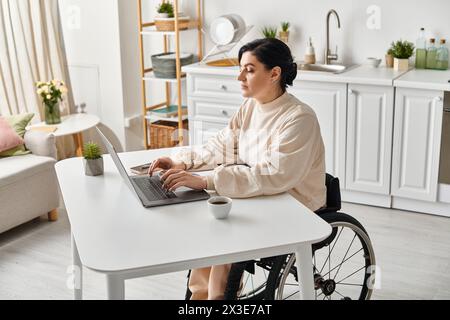 Une femme handicapée en fauteuil roulant travaille à distance sur son ordinateur portable dans la cuisine, mettant en valeur son indépendance numérique. Banque D'Images