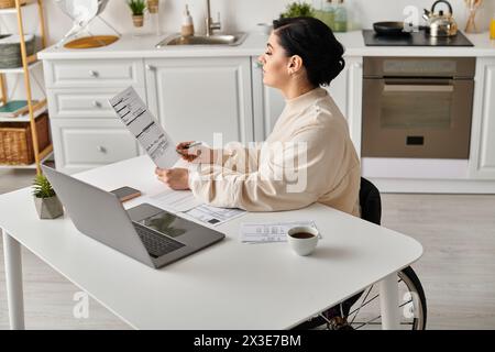 Une femme handicapée en fauteuil roulant travaille à distance depuis sa cuisine, concentrée sur son ordinateur portable sur la table. Banque D'Images