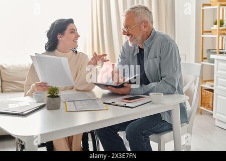 Un homme et une femme handicapée en fauteuil roulant sont assis à une table, engagés dans une conversation profonde dans leur cuisine à la maison. Banque D'Images