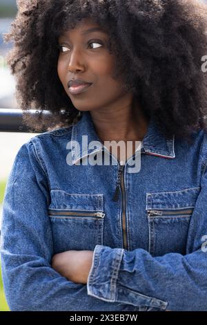 Un gros plan représente une femme noire avec un afro, vêtue d'une veste en denim classique, regardant avec une expression rêveuse. La mise au point douce sur son visage renforce son regard contemplatif, suggérant une pensée profonde ou une aspiration, dans un contexte urbain. Dreamy Black femme en veste denim regardant loin. Photo de haute qualité Banque D'Images