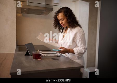 Jeune jolie femme à la maison, tient une facture papier ou une lettre à l'aide d'une tablette numérique et d'un smartphone, effectuant des paiements en ligne sur le site Web, calculant le t financier Banque D'Images