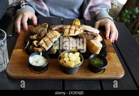 Plat de gril mixte comprend du poulet, du steak et des légumes, à côté de deux types de sauces et frites, une femme dînant en plein air Banque D'Images