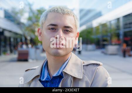 portrait d'un jeune homme gay aux cheveux blonds courts, calme dans la rue debout à l'extérieur des bureaux de travail regardant calmement la caméra, portant une veste robe marron an Banque D'Images
