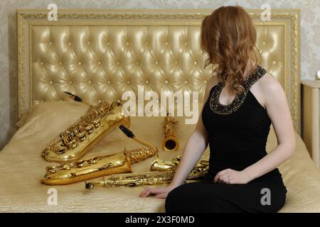 Femme en robe noire assise avec sax sur le lit avec des instruments à vent en studio Banque D'Images