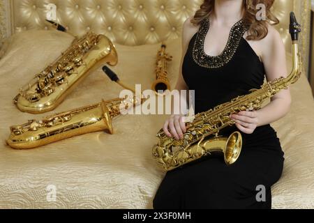 Femme en robe noire assise avec sax sur le lit avec des instruments à vent, noface Banque D'Images