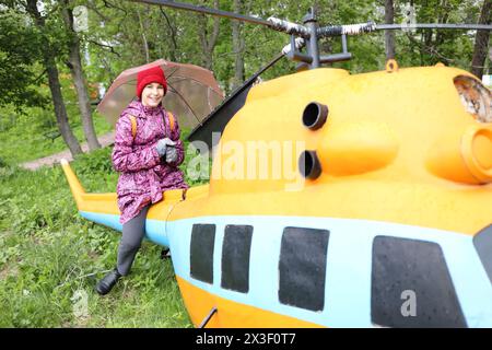 Fille souriante avec parapluie se trouve sur un petit hélicoptère jaune vert dans le parc Banque D'Images