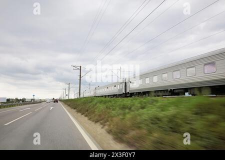 Le train se déplace sur le chemin de fer et les voitures se déplacent sur l'autoroute à la journée nuageuse d'été Banque D'Images