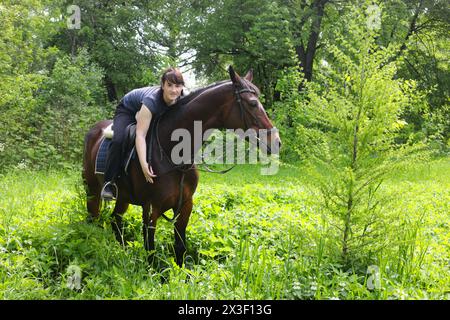 Jolie fille heureuse est assise sur le cheval parmi les arbres dans la forêt verte d'été Banque D'Images