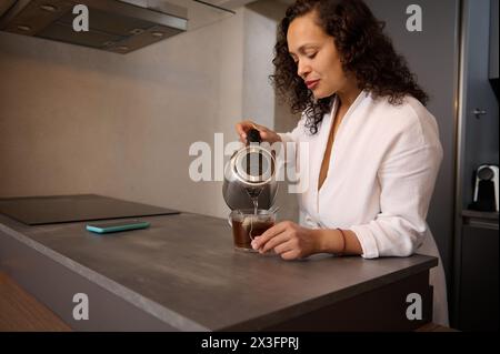 Jeune femme brune multi ethnique aux cheveux bouclés faisant du thé pour le petit déjeuner. Jolie dame sur un peignoir blanc versant de l'eau bouillie d'une théière électrique Banque D'Images
