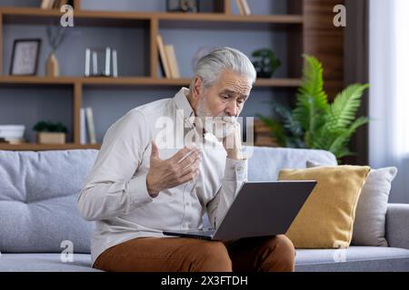 Homme âgé aux cheveux gris bouleversé parlant sur un appel vidéo avec la famille et la banque, assis sur le canapé et résolvant des problèmes et des questions financières. Banque D'Images