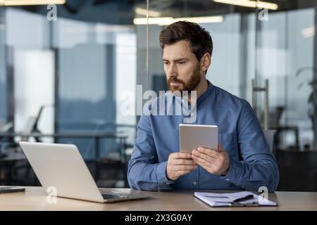 Un jeune homme sérieux travaille au bureau à un bureau, tient une couverture et regarde attentivement l'écran d'un ordinateur portable, compare les données et fait une analyse. Banque D'Images