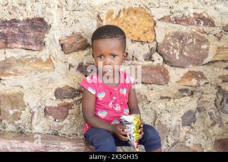 fille africaine du village mangeant des collations, situé dans la cour en face de la maison Banque D'Images