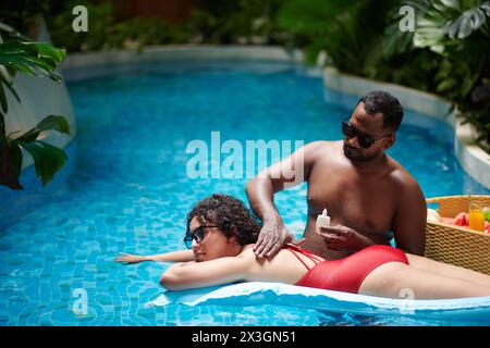 Jeune femme se relaxant sur matelas d'air sur la surface de l'eau bleue dans la piscine tandis que son petit ami appliquant la lotion sur son dos Banque D'Images