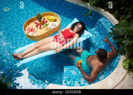 Jeune couple reposant passant du temps dans la piscine avec de l'eau bleue transparente tandis que l'homme ayant boire et la femme se relaxant sur le matelas d'air Banque D'Images