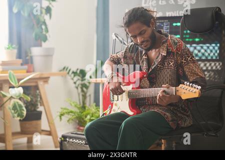 Jeune homme talentueux d'origine indienne assis devant la caméra dans le home studio et jouant de la guitare électrique tout en créant de la nouvelle musique Banque D'Images