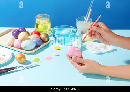 Beaucoup d'œufs de Pâques ont été peints exposés sur un plateau, disposés avec un bol et des tasses d'eau, des pinceaux, des couleurs de peinture. Le modèle de main tient un paintbrus Banque D'Images