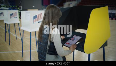 ZAPORIZHYA, UKRAINE -15 MARS 2024 : femme caucasienne, électrice pense et fait un choix debout près de l'isoloir au bureau de vote. Un citoyen AMÉRICAIN utilise une tablette pour voter. Election présidentielle aux États-Unis d'Amérique. Banque D'Images