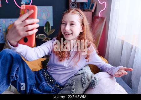 Adolescente joyeuse tenant un smartphone assis sur son lit discutant lors d'un appel vidéo, vue en angle élevé Banque D'Images