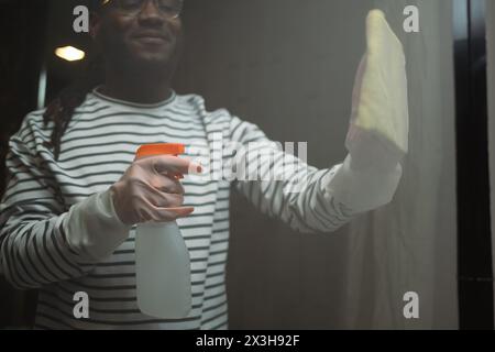 Homme africain souriant nettoyant le verre de douche dans la salle de bains avec spray de nettoyage et chiffon Banque D'Images