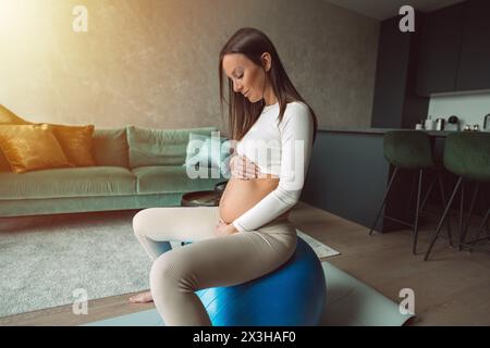 Calme femme enceinte assise sur un ballon de fitness et touchant son ventre. Mère heureuse faisant des exercices physiques de soutien pour une grossesse en douceur. Banque D'Images