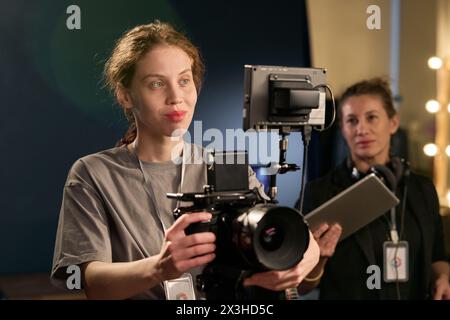 Portrait de taille haute de jeune femme souriante exploitant un équipement de caméra professionnel sur le plateau pendant l'espace de copie de production vidéo Banque D'Images