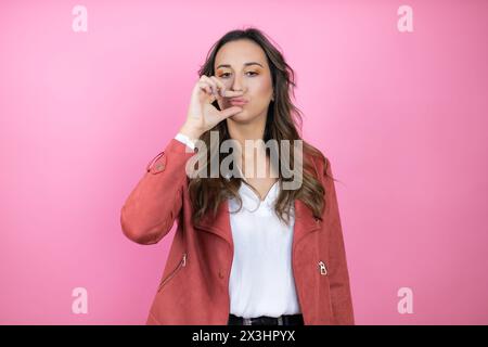 Jeune belle femme portant une veste décontractée sur fond rose isolé bouche et lèvres fermées comme zip avec les doigts. Secret et silencieux, parler tabou Banque D'Images