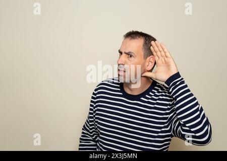 Homme hispanique à barbe dans ses années 40 portant un pull rayé écoutant des rumeurs garder la main près de l'oreille isolée sur fond de couleur beige. Banque D'Images