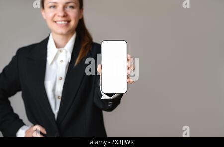 Femme d'affaires en costume montre l'écran de son smartphone à la caméra et souriant. Maquette d'écran blanc de téléphone portable dans la main de femme d'affaires Banque D'Images