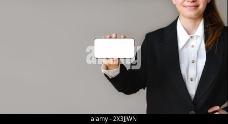 Maquette de l'écran blanc du téléphone portable dans la main de la femme d'affaires sur fond gris. Femme d'affaires en costume montre l'écran de son smartphone t Banque D'Images