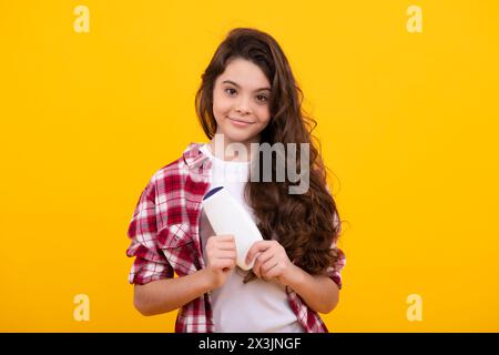 Jeune fille avec flacon de shampooing ou gel douche isolé sur fond jaune. Produit cosmétique pour les cheveux pour enfants. Joyeux adolescent, positif et souriant emot Banque D'Images