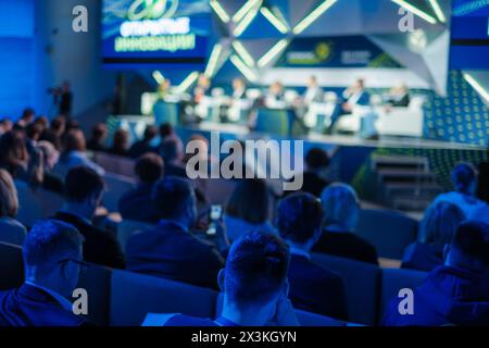 Les membres du public vêtus d'une tenue formelle se concentrent lors d'une conférence d'affaires animée et participent à des discussions dans un lieu moderne éclairé par un éclairage bleu dynamique. Banque D'Images